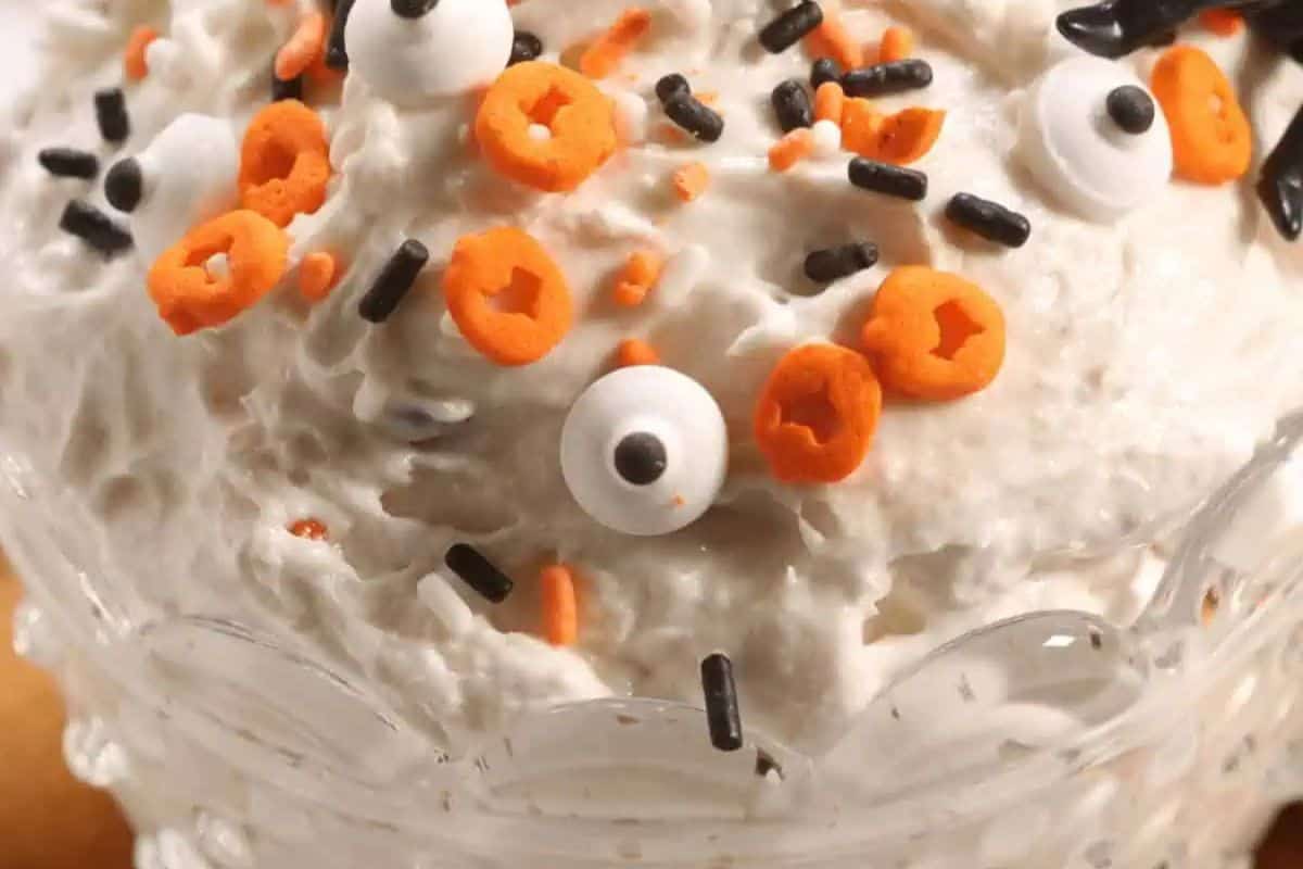 A close-up of a clear bowl filled with a white, fluffy dip. It's decorated with Halloween sprinkles, including orange pumpkin shapes, black and white jimmies, and a single candy eyeball.
