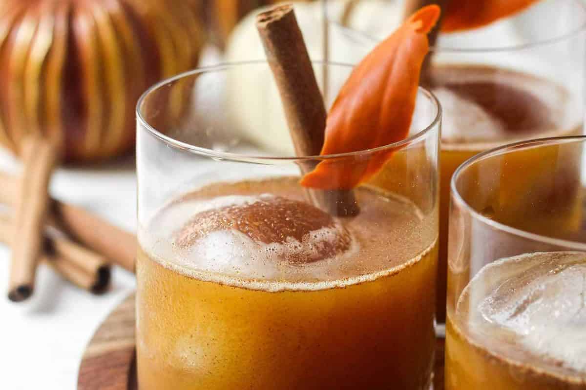 A glass of brown spiced drink with a large ice cube, garnished with a cinnamon stick and an orange leaf—perfect inspiration for autumn cocktails. Other glasses and cinnamon sticks are in the background.