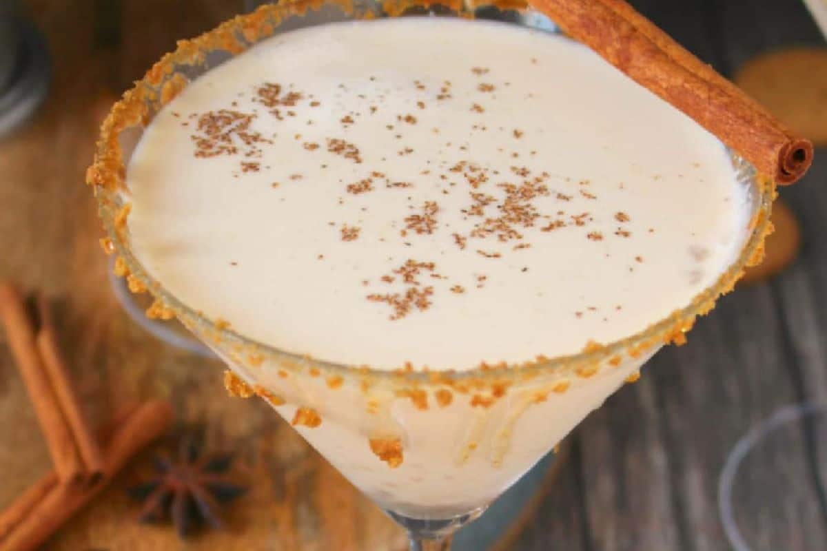 A creamy autumn cocktail in a martini glass with a crumb-coated rim, topped with grated spice and garnished with a cinnamon stick—perfect for fans of cozy fall drinks.