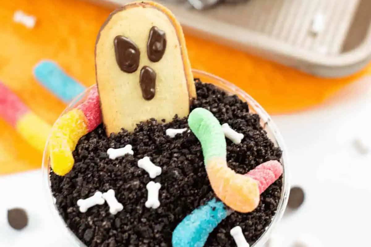 A small dessert cup filled with chocolate cookie crumbs to look like dirt. A gingerbread-man-shaped cookie with a ghost face is sticking out of the "dirt," along with gummy worms and small white bone-shaped candies.
