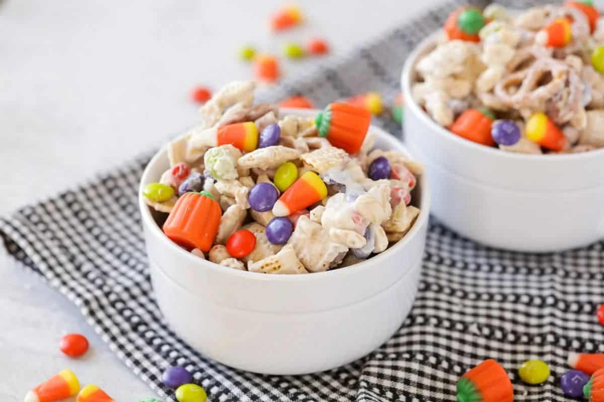 A white bowl filled with a Halloween-themed snack mix. The mix includes white-chocolate coated cereal and pretzels, candy corn, M&Ms, and pumpkin-shaped candies. A similar bowl is visible in the background, all on a black and white checkered napkin.