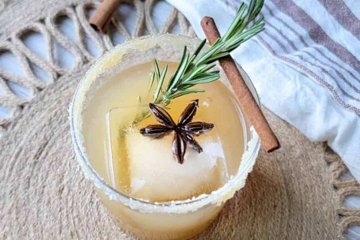 A cocktail with a large ice cube, garnished with star anise and a rosemary sprig, sits on a woven mat next to a cinnamon stick and striped cloth—perfect for fans of fall drinks and cozy Autumn cocktails.