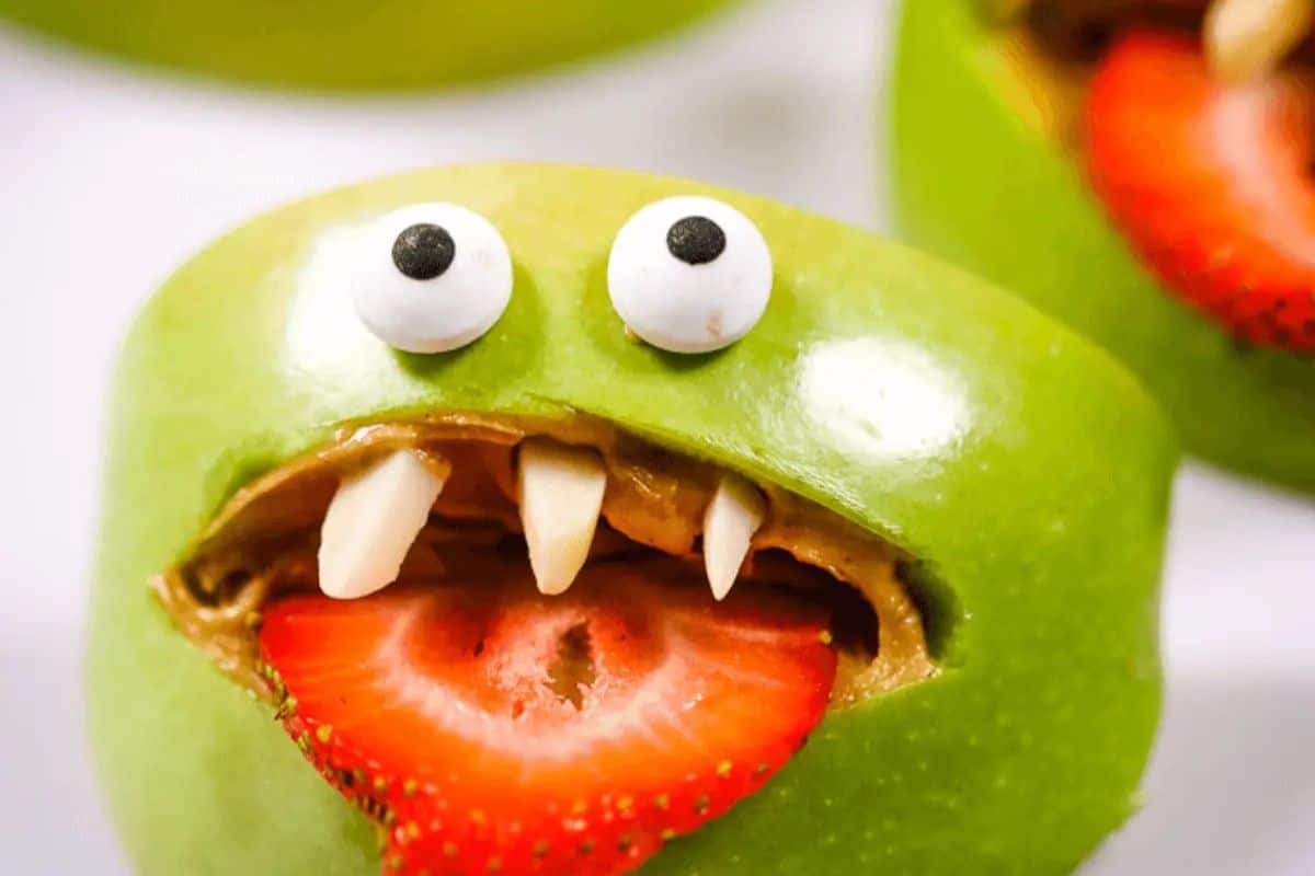  A close-up of a green apple carved to look like a monster mouth. It's filled with a nut butter, has sliced almonds for teeth, candy googly eyes, and a slice of strawberry sticking out like a tongue.