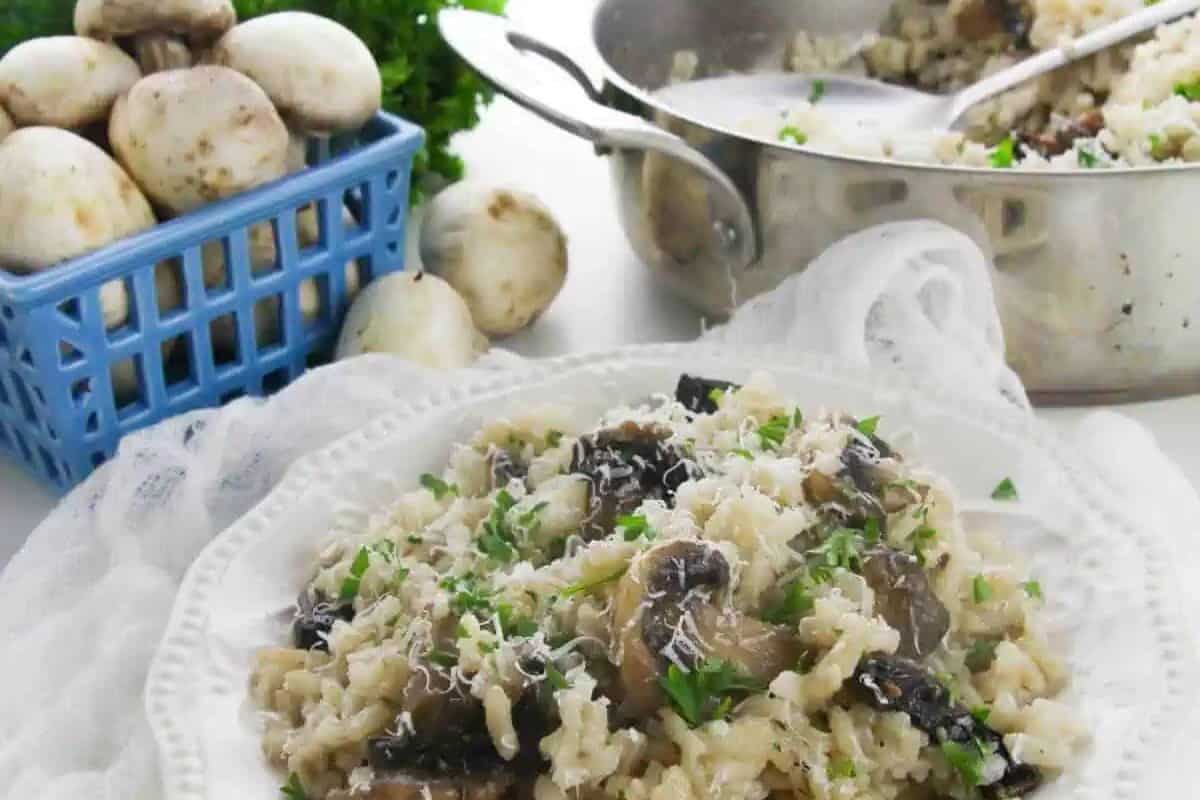 A plate of mushroom risotto garnished with grated cheese and parsley, with a pan of risotto and fresh mushrooms in the background—a perfect comfort food for cozy evenings and comforting fall meals.