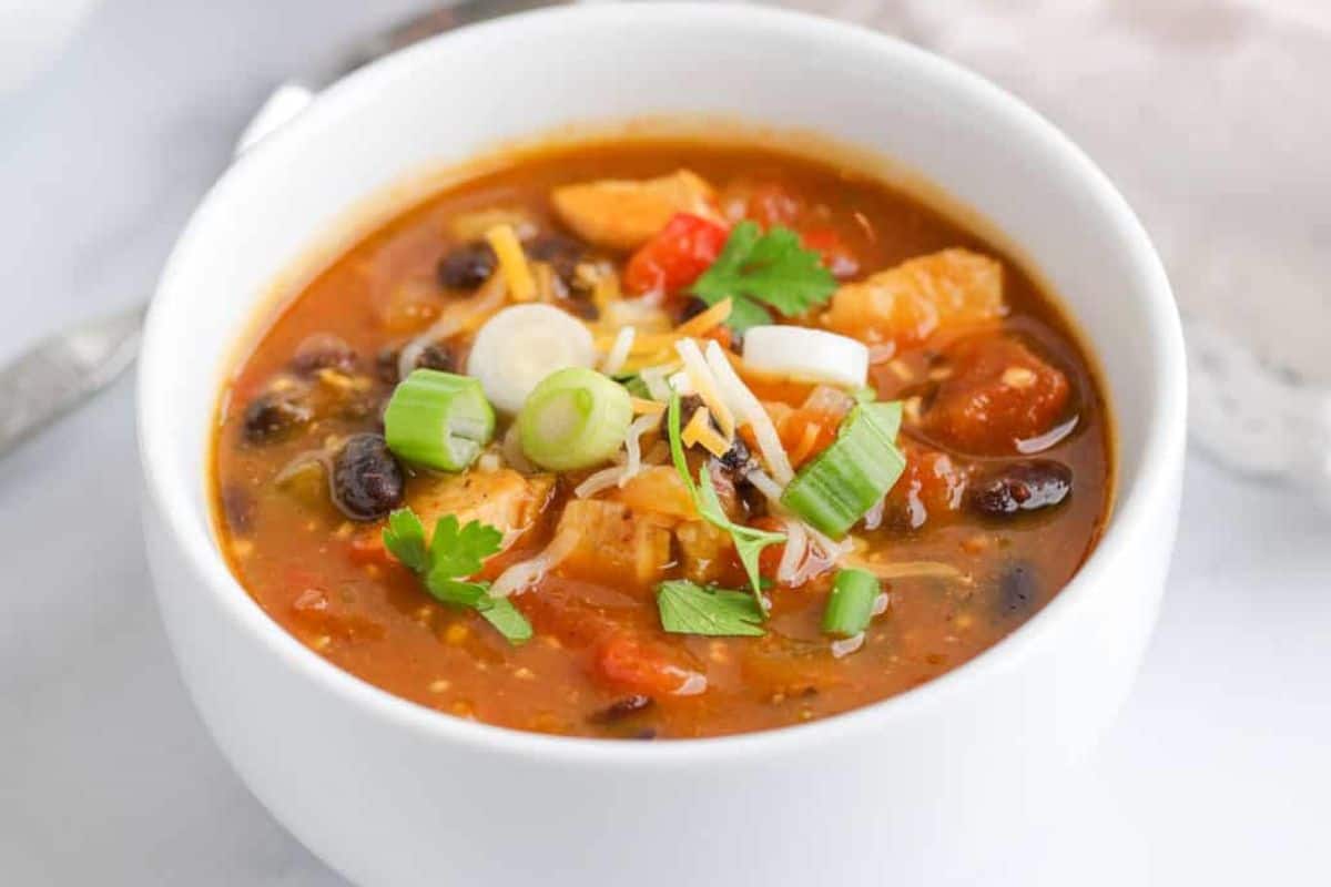 A white bowl filled with chunky soup containing beans, vegetables, cheese, and topped with sliced green onions and fresh herbs—perfect comfort food for those seeking comforting fall meals.