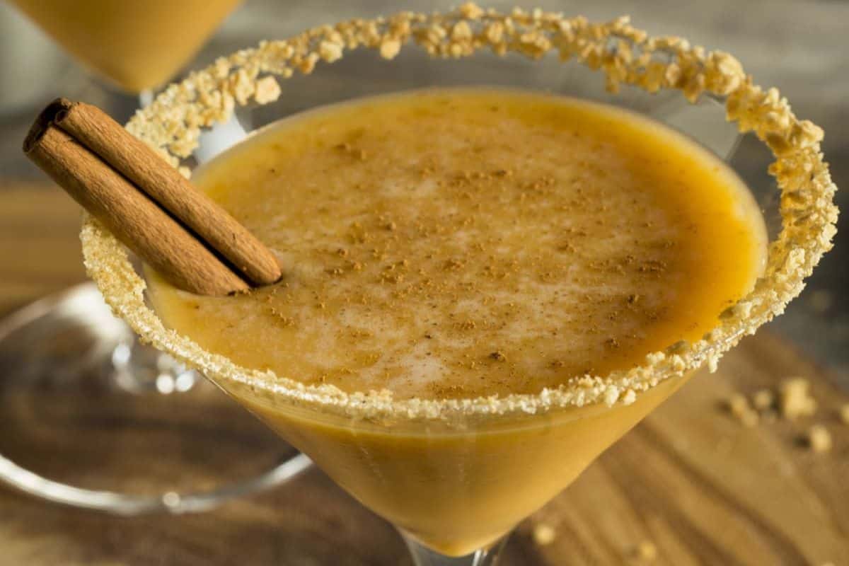 A pumpkin spice cocktail in a martini glass with a crumb-coated rim, topped with a cinnamon stick—a cozy favorite among fall drinks.
