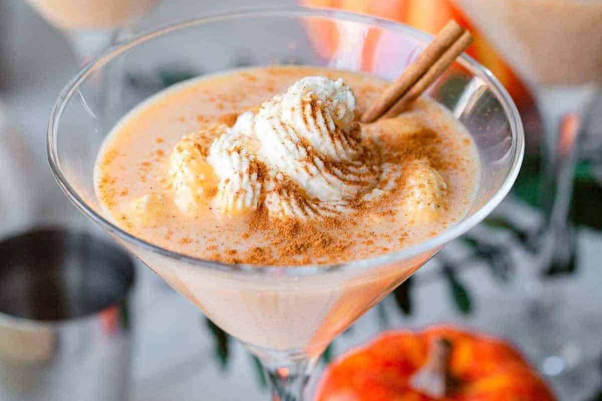 A close-up of a creamy orange drink in a martini glass, topped with whipped cream, ground cinnamon, and a cinnamon stick—perfect for those who love seasonal cocktails or cozy autumn cocktails.