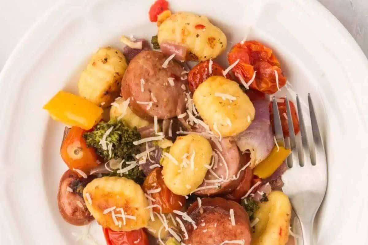 A white plate with gnocchi, roasted potatoes, cherry tomatoes, broccoli, bell peppers, onions, and shredded cheese offers cozy comfort food perfect for fall meals. A fork sits on the side, ready for you to enjoy this seasonal recipe.