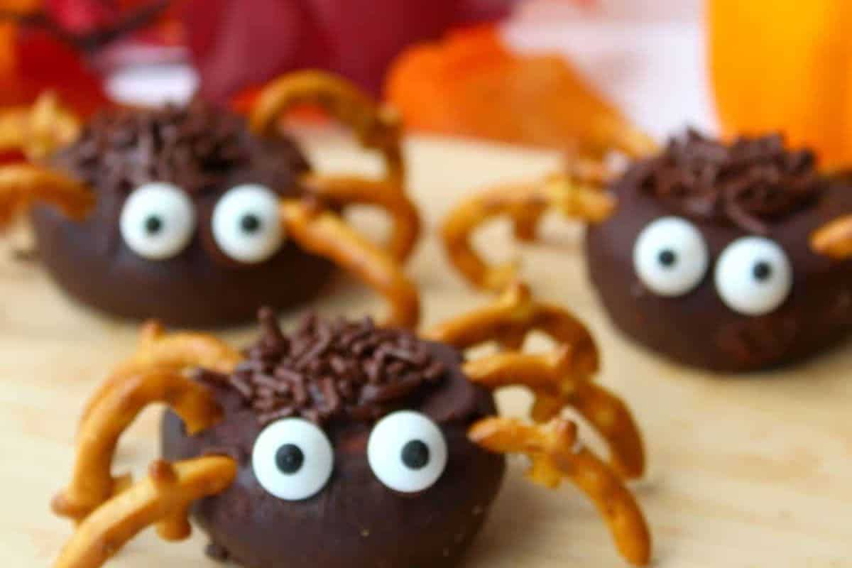 Three chocolate donuts decorated to look like spiders. They have brown sprinkles for hair, candy googly eyes, and pretzel legs sticking out from the sides.