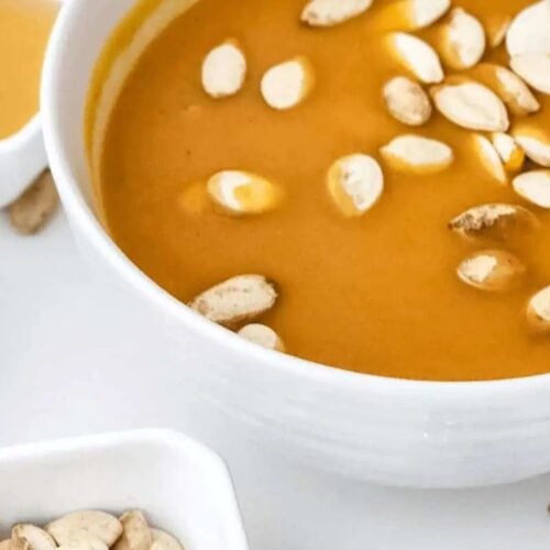 A bowl of creamy orange soup topped with pumpkin seeds, surrounded by small dishes containing spices, seeds, and sauce on a white surface—perfect for comforting recipes and cozy Fall meals.