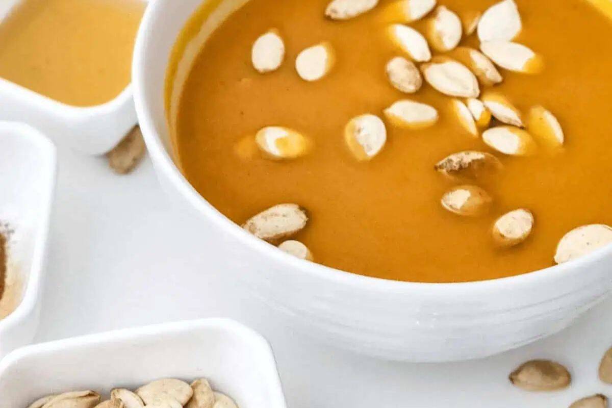 A bowl of creamy orange soup topped with pumpkin seeds, surrounded by small dishes containing spices, seeds, and sauce on a white surface—perfect for comforting recipes and cozy Fall meals.