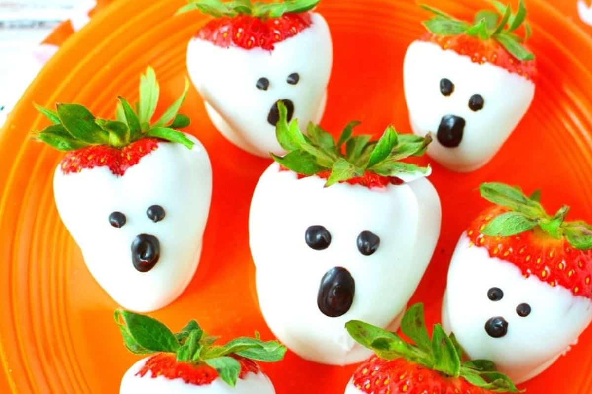 Strawberries dipped in white chocolate with small dark chocolate dots for ghost eyes, resembling spooky ghost shapes.