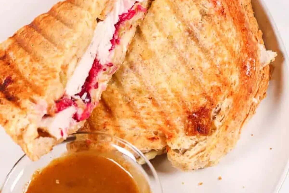 A toasted sandwich with turkey and cranberry filling, perfect for Fall meals, is served on a plate with a small bowl of brown dipping sauce.