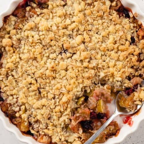 A round dish of Apple Berry Crisp with a golden oat topping, partially served with a metal spoon resting in the dish.