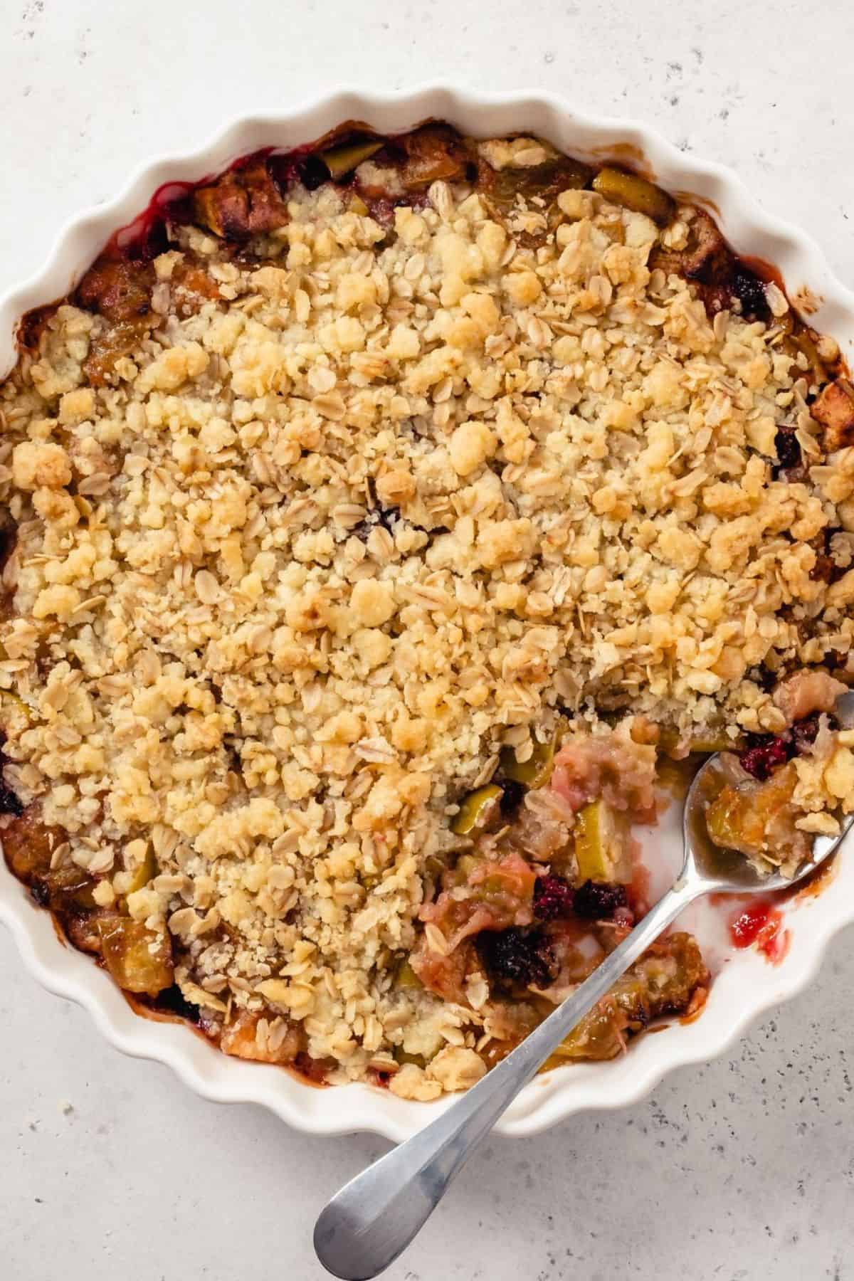 A round dish of Apple Berry Crisp with a golden oat topping, partially served with a metal spoon resting in the dish.