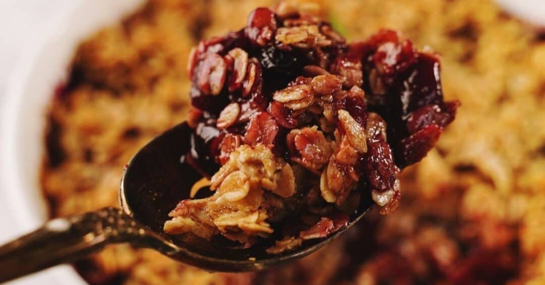 A close-up of a spoon holding a serving of baked Apple Berry Crisp, with the rest of the dish blurred in the background.