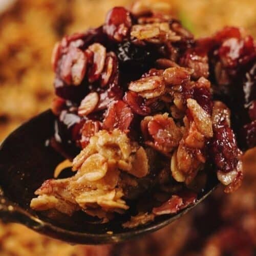A close-up of a spoon holding a serving of baked Apple Berry Crisp, with the rest of the dish blurred in the background.