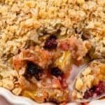 A close-up of an Apple Berry Crisp in a white dish, showing a crumbly oat topping and a spoonful revealing the juicy berry and fruit filling underneath.