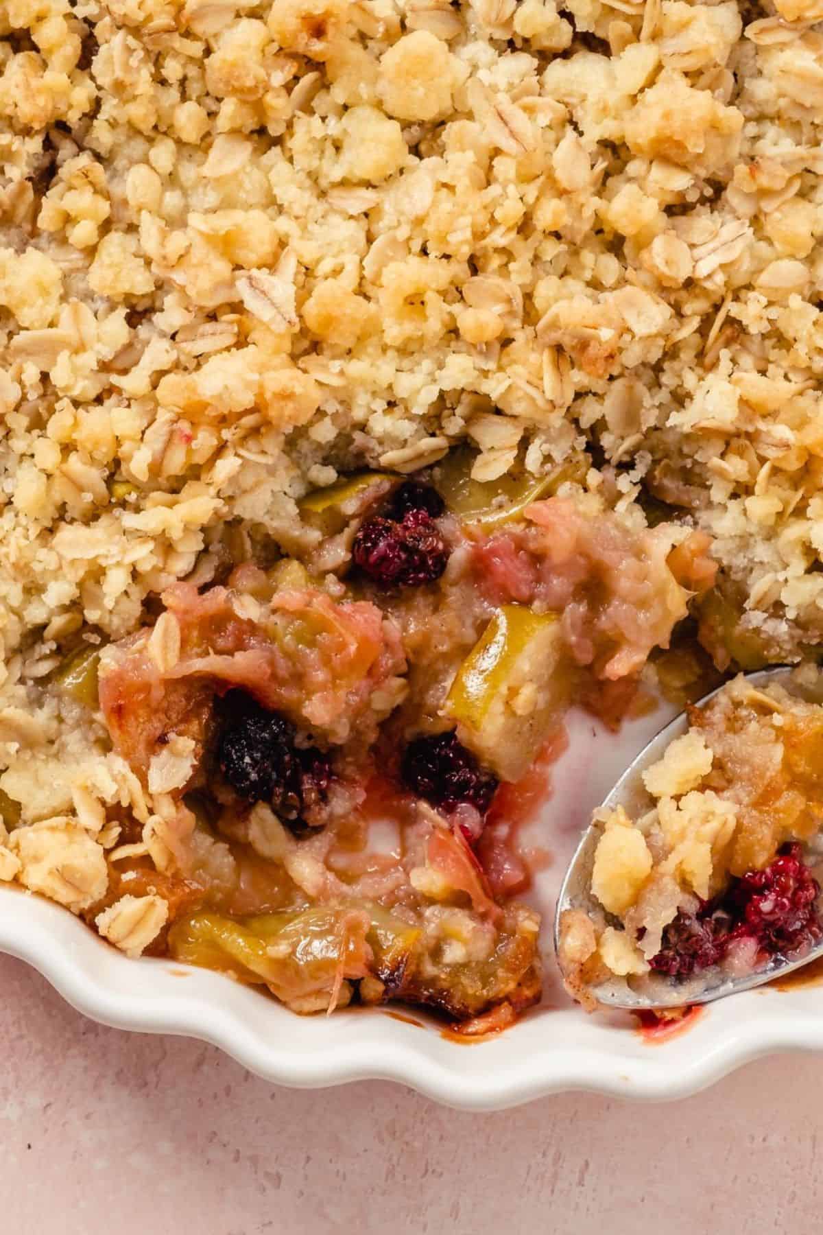 A close-up of an Apple Berry Crisp in a white dish, showing a crumbly oat topping and a spoonful revealing the juicy berry and fruit filling underneath.