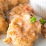Close-up of Bang Bang Shrimp, breaded and covered in a creamy sauce, garnished with chopped green onions, served on a white plate with a dipping sauce in the background.