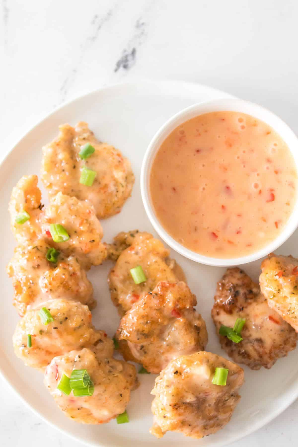 A plate of breaded, sauced nuggets inspired by Bang Bang Shrimp, garnished with chopped green onions and served next to a bowl of creamy orange dipping sauce.