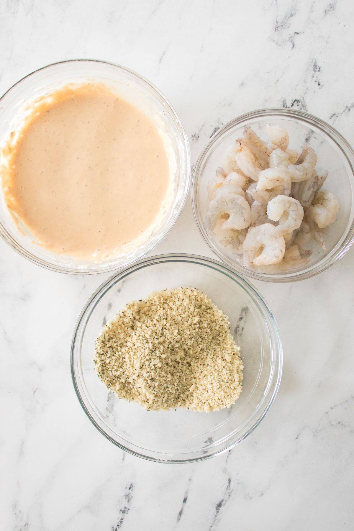 Three glass bowls for Bang Bang Shrimp—one with batter, one with raw shrimp, and one with breadcrumbs—are arranged on a white marble surface.