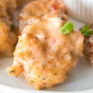 A close-up of a battered and sauced piece of fried chicken garnished with chopped green onion on a white plate, reminiscent of the spicy-sweet flavors found in Bang Bang Shrimp.