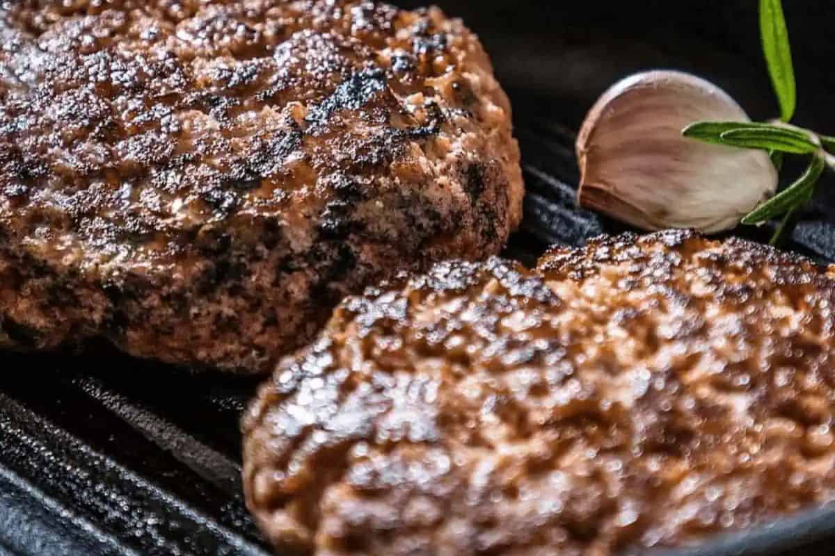Two browned beef burger patties sizzle on a grill beside a clove of garlic and rosemary—a classic choice for cast iron recipes.