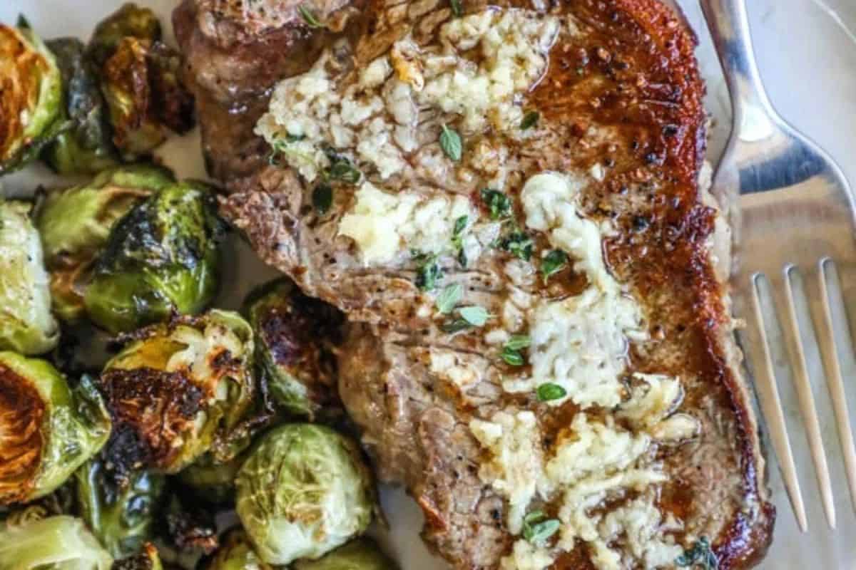 A cooked steak from classic cast iron recipes, topped with melting butter and herbs, is served next to roasted Brussels sprouts on a white plate with a fork.