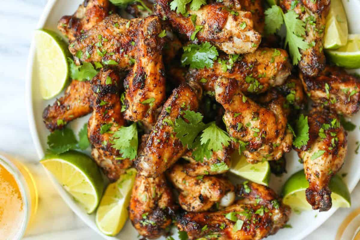 A plate of grilled chicken wings garnished with cilantro, surrounded by lime wedges. A glass of beer is partially visible on the side.