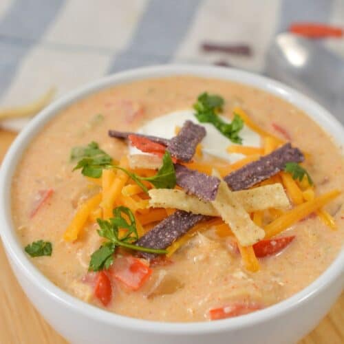 A bowl of creamy Crock Pot Chicken Chili soup topped with shredded cheese, tortilla strips, sour cream, and chopped herbs, placed on a wooden surface.