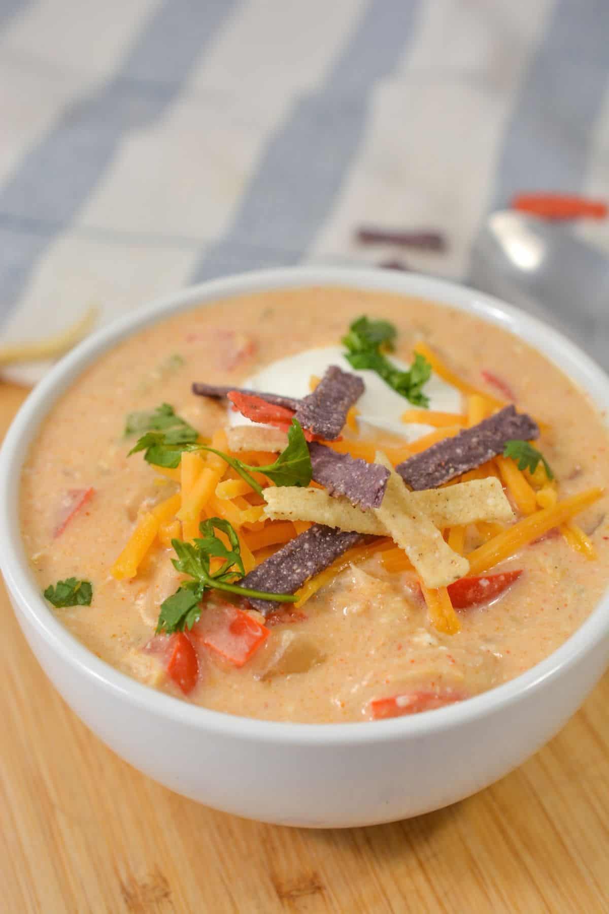 A bowl of creamy Crock Pot Chicken Chili soup topped with shredded cheese, tortilla strips, sour cream, and chopped herbs, placed on a wooden surface.
