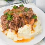 A serving of Crockpot Short Ribs Braised in Beer, shredded with gravy and chopped parsley on top of mashed potatoes on a white plate.