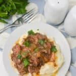 A plate of mashed potatoes topped with Crockpot Short Ribs braised in beer and garnished with chopped parsley, next to a fork, salt and pepper shakers, and fresh parsley.