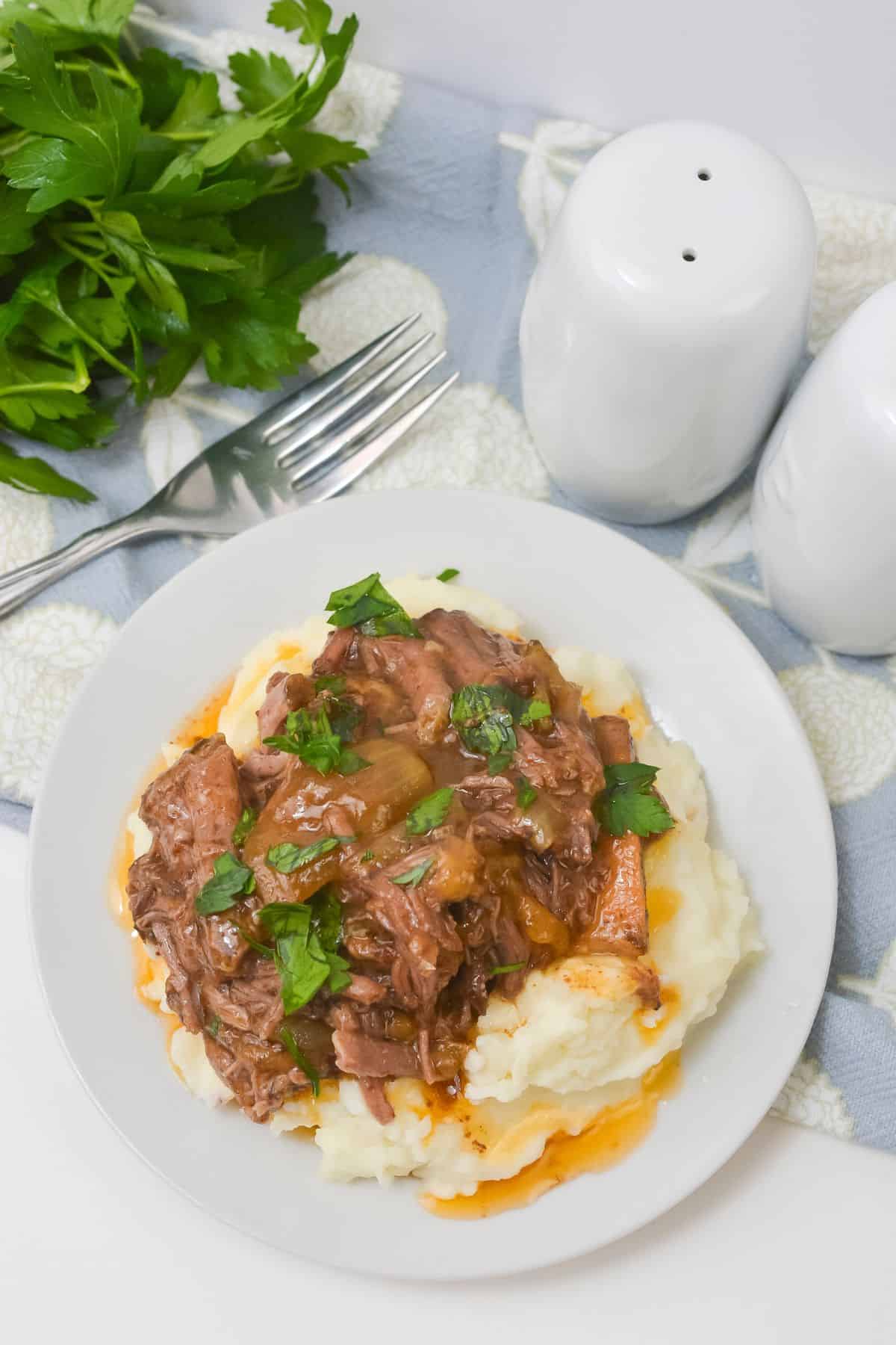 A plate of mashed potatoes topped with Crockpot Short Ribs braised in beer and garnished with chopped parsley, next to a fork, salt and pepper shakers, and fresh parsley.