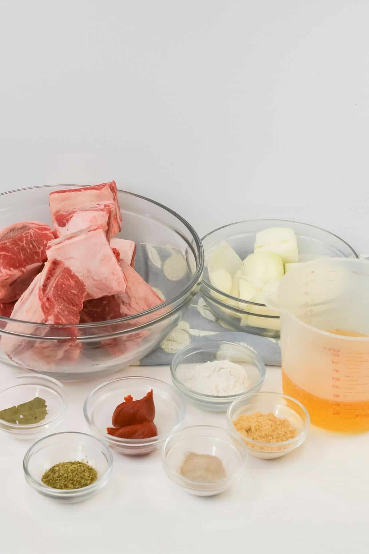 Raw beef bones, chopped onions, minced garlic, and various seasonings and liquids are arranged in separate bowls—perfect prep for Crockpot Short Ribs Braised in Beer—on a clean white surface.