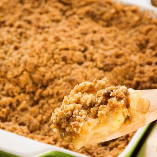 A close-up of a wooden spoon holding a serving of Easy Apple Crumble with crumb topping, in front of a baking dish filled with more apple crisp.