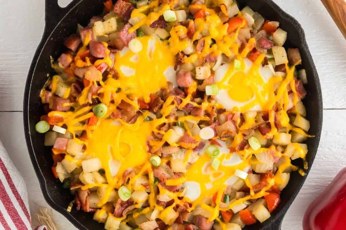 A cast iron skillet filled with hash made of diced potatoes, ham, bell peppers, melted cheese, green onions, and two eggs—perfect for those seeking delicious breakfast recipes.