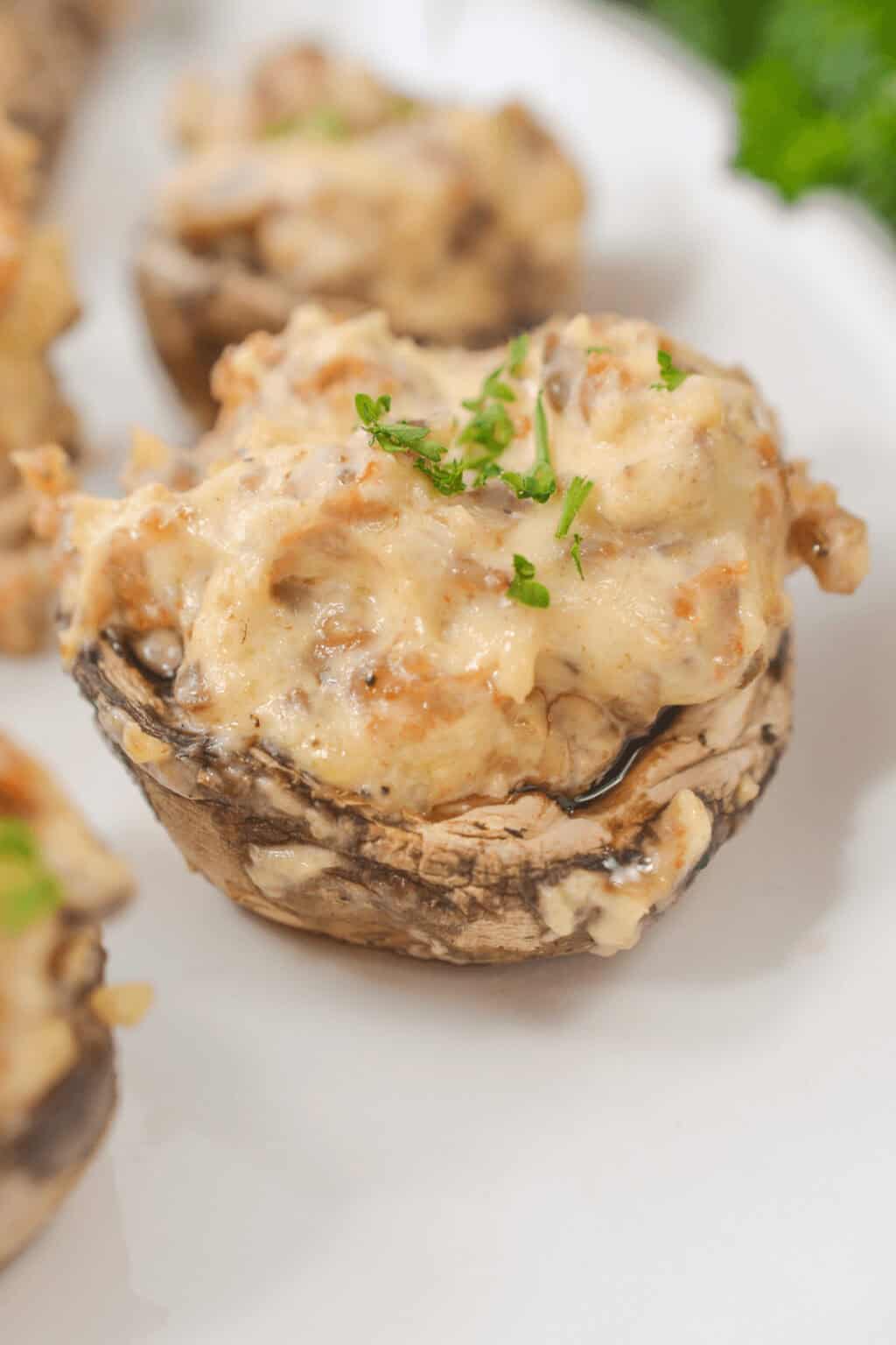 Cream cheese filled mushrooms are delicately topped with a creamy filling and garnished with chopped parsley, all elegantly arranged on a white plate.