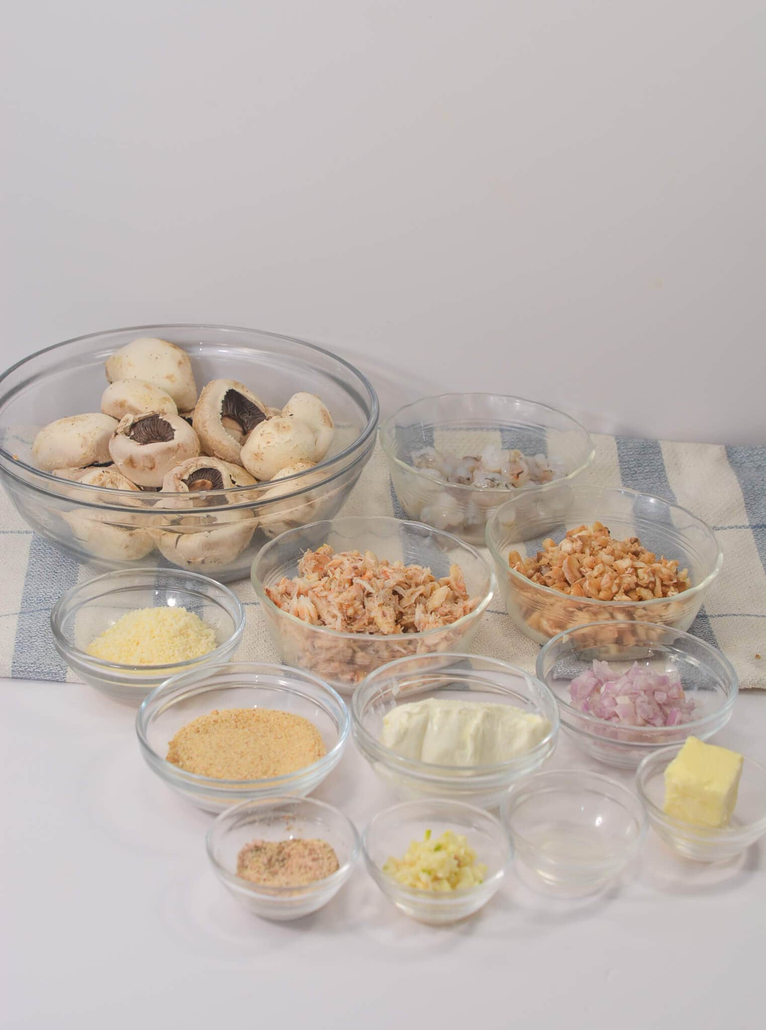 An arrangement of small bowls featuring crab-stuffed portobello mushrooms alongside prawns, chopped nuts, cheese, breadcrumbs, butter, garlic, onions, and cream rests on a tablecloth.