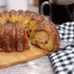 A Bundt coffee cake with a slice cut out rests on a wooden board, complemented by a checkered cloth and a steaming cup of black coffee.