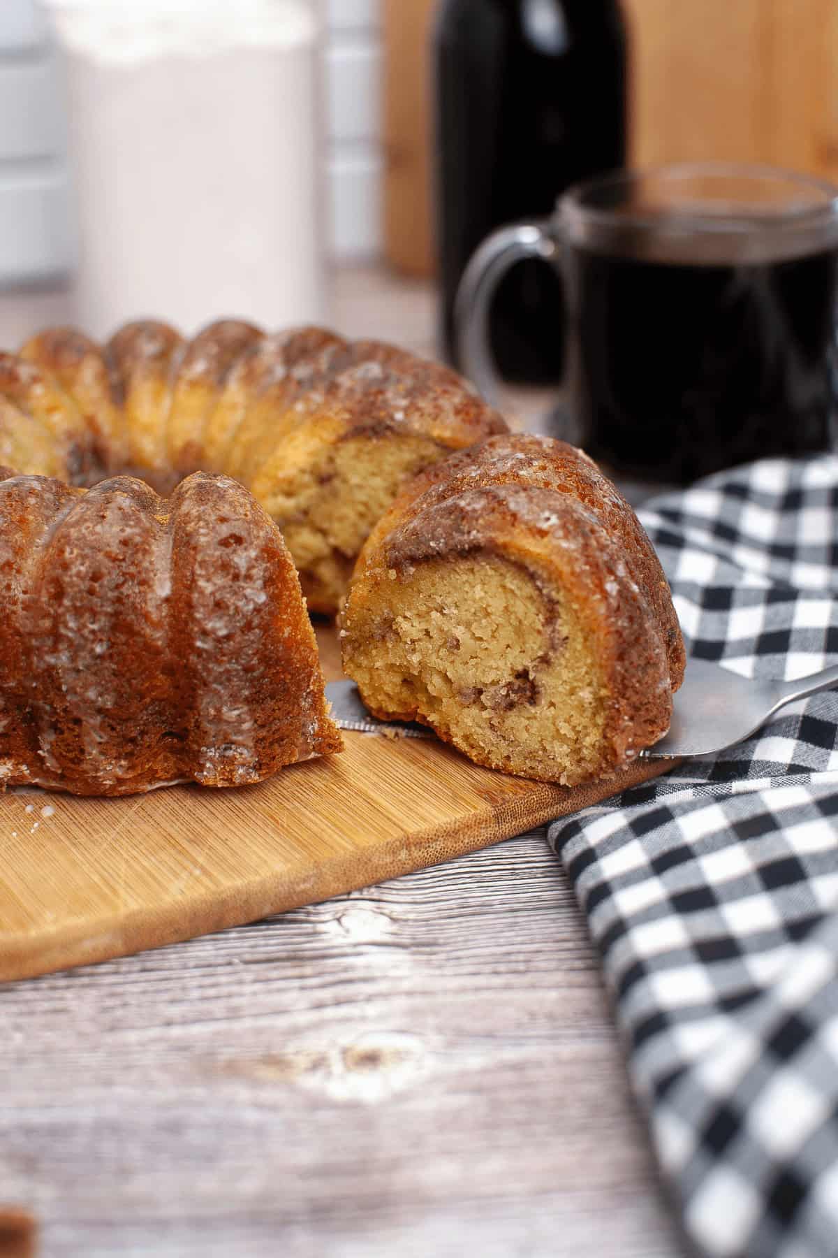 A Bundt coffee cake with a slice cut out rests on a wooden board, complemented by a checkered cloth and a steaming cup of black coffee.
