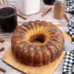 A tempting bundt coffee cake rests on a wooden board with a knife beside it. Nearby, measuring cups and cinnamon sticks hint at the rich flavors within, while a glass of dark liquid and a black-and-white checkered cloth complete the cozy scene.