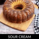 A rich bundt coffee cake with sour cream rests on a wooden board, accompanied by a knife and a checkered cloth.