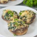 Three stuffed mushrooms filled with spinach, Boursin cheese, and herbs are arranged on a white plate, with fresh parsley in the background.