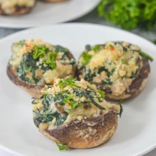 Three stuffed mushrooms filled with spinach, Boursin cheese, and herbs are arranged on a white plate, with fresh parsley in the background.