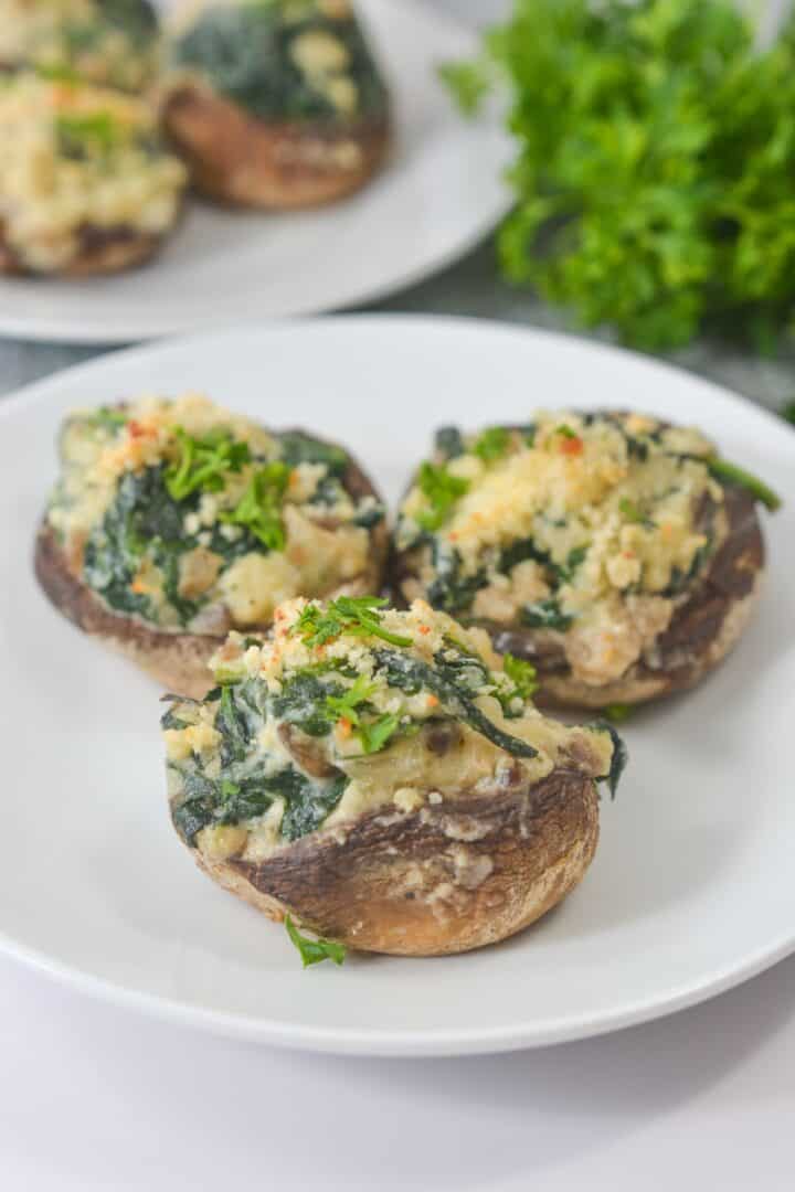 Three stuffed mushrooms filled with spinach, Boursin cheese, and herbs are arranged on a white plate, with fresh parsley in the background.