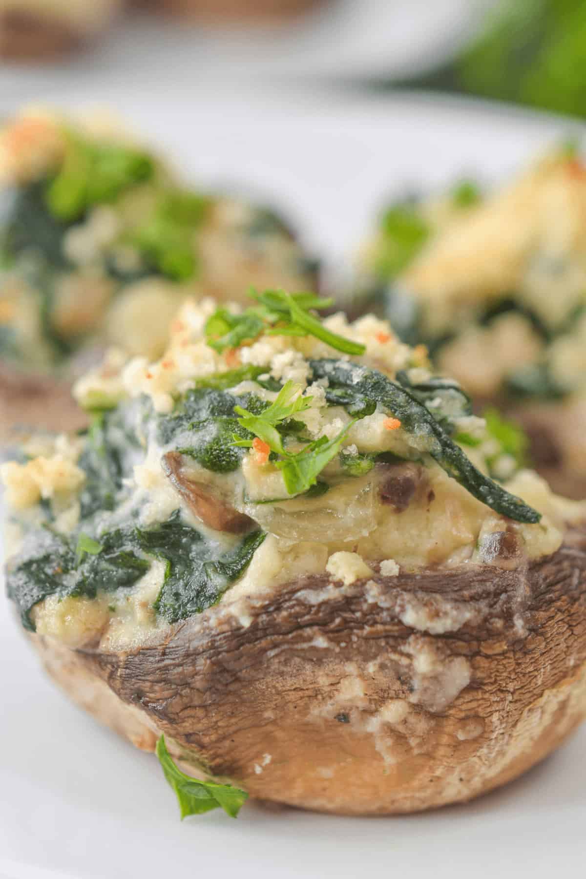 A close-up of a boursin cheese stuffed mushroom cap topped with spinach, breadcrumbs, and herbs sits elegantly on a white plate.