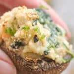 A hand holds a stuffed mushroom with a creamy Boursin cheese filling, topped with fresh herbs.