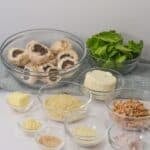 Various clear bowls containing butter, breadcrumbs, grated cheese, minced garlic, chopped nuts, and chopped onions arranged on a white surface.